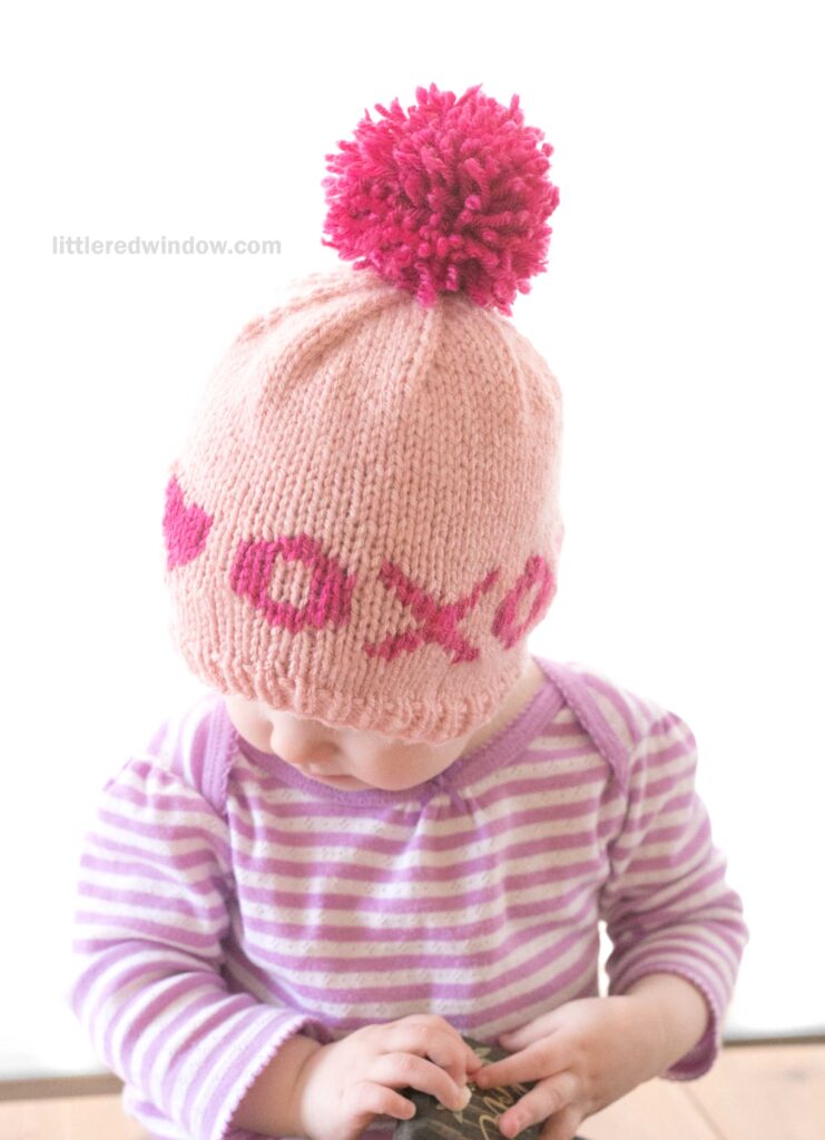 A baby wearing a pink valentine hat with a knitted pom-pom and the letters